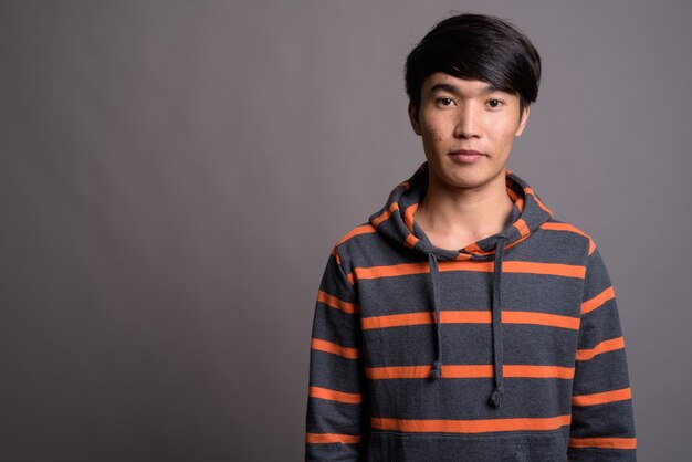 Junger asiatischer Mann, der gestreiften Kapuzenpulli gegen graue Wand trägt