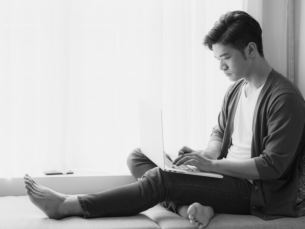 Junger asiatischer Mann, der in einem Buch schreibt, wenn er Laptop benutzt