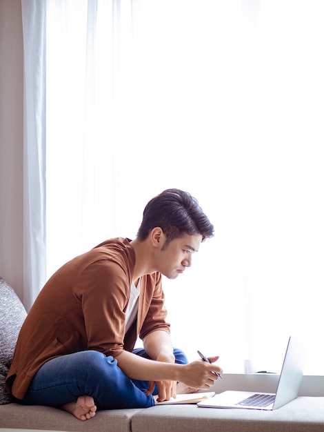 Junger asiatischer Mann, der in Notizbuch schreibt und Laptop verwendet.