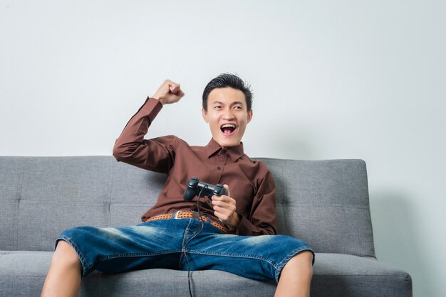 Junger asiatischer Mann, der Joystick für das Spielen des Fußballvideospiels beim Sitzen auf Sofa im Wohnzimmer hält.