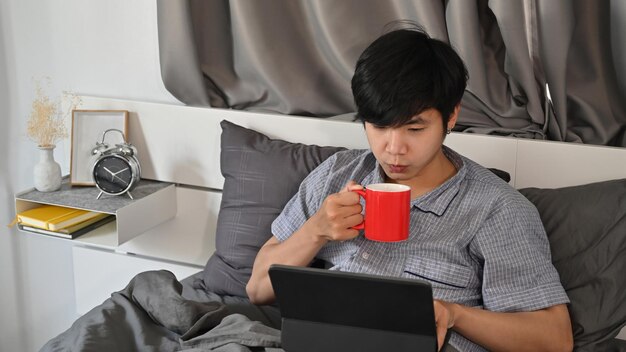 Junger asiatischer Mann, der Kaffee trinkt und Computertablette auf seinem Bett verwendet.