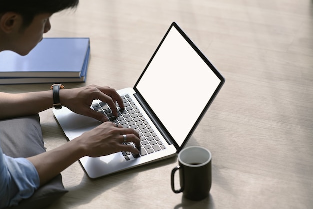 Junger asiatischer Mann, der mit Computer-Laptop arbeitet, während auf Holzboden im Wohnzimmer zu Hause liegend.