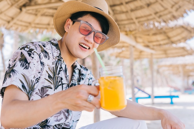 Junger asiatischer Mann, der Orangensaft am Strand trinkt