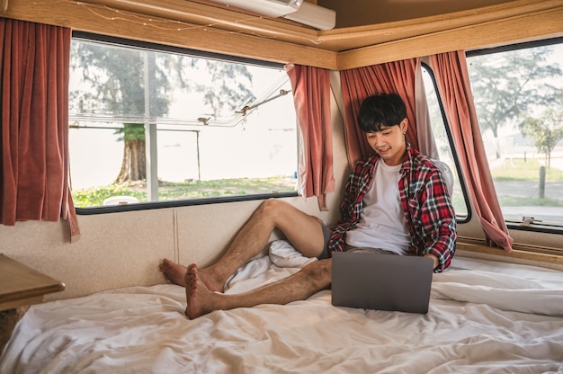 Junger asiatischer Mann, der schottisches Hemd trägt, das mit Laptop auf dem Bett im Wohnmobil entspannt