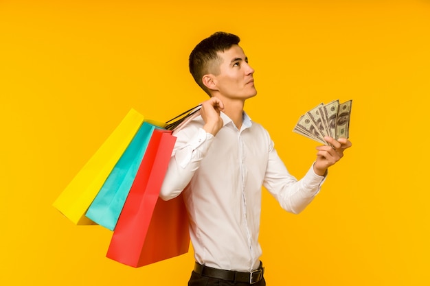 Junger asiatischer Mann, der seine Einkaufstasche und Geld auf gelbem Hintergrund zeigt