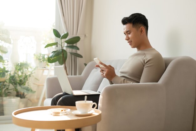 Junger asiatischer Mann, der sich zu Hause auf einem Sofa mit seinem Laptop entspannt, der auf seinem Handy chattet, während er mit einem Lächeln den Bildschirm des Laptops liest