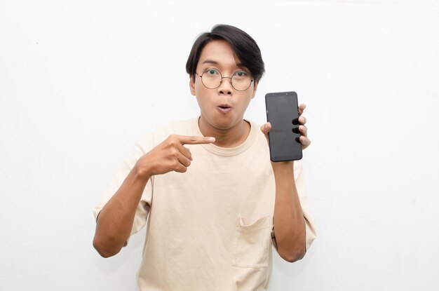 junger asiatischer mann im beigen t-shirt, das telefonbildschirm mit glücklichem euphorischem schockiertem gesicht hält und zeigt