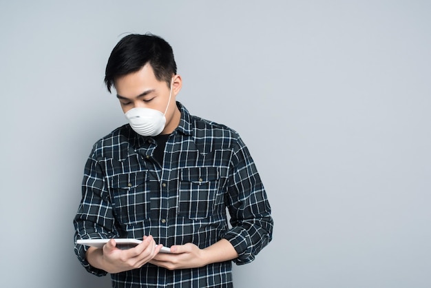 Junger asiatischer Mann in Atemschutzmaske mit digitalem Tablet auf grauem Hintergrund