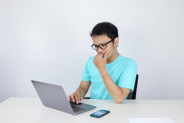 Junger asiatischer Mann ist ernst und konzentriert sich, wenn er an einem Laptop auf dem Tisch arbeitet Indonesischer Mann mit blauem Hemd