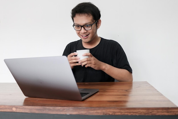 Junger asiatischer Mann lächelt und genießt es, wenn er zu Hause mit Laptop und einer Tasse Kaffee arbeitet Ein Mann sitzt auf dem Bett und arbeitet Arbeiten von zu Hause aus Konzept