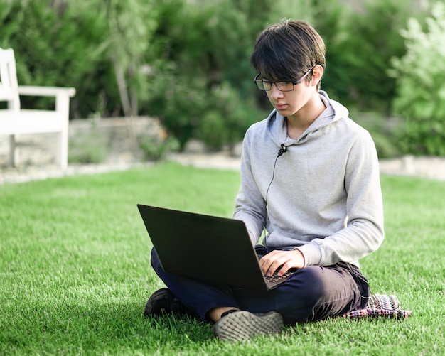 Junger asiatischer Mann mit Brille arbeiten am Laptop auf dem Rasen im Freien