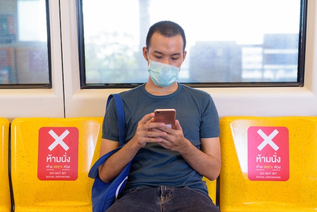 Junger asiatischer Mann mit Maske unter Verwendung des Telefons und sitzend mit Abstand innerhalb des Zuges