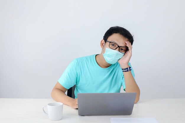 Junger asiatischer Mann mit medizinischer Maske fühlt sich ungesund müde und verwirrt mit der Arbeit am Laptop