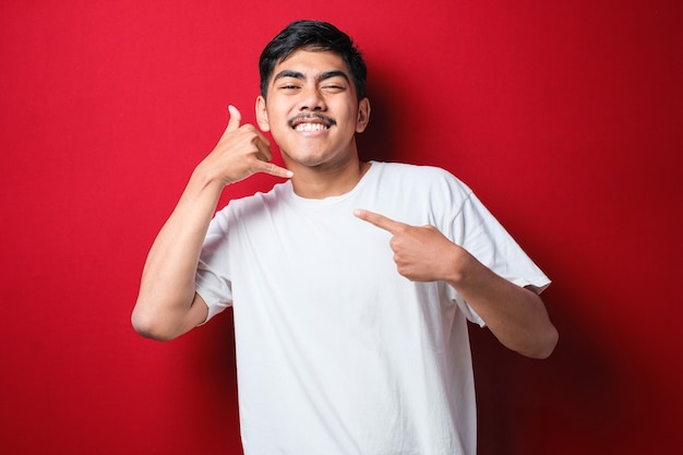 Junger asiatischer Mann mit weißem T-Shirt, der in die Kamera lächelt und zeigt, rufen Sie mich an, Telefon-Handzeichen-Geste, vor rotem Hintergrund