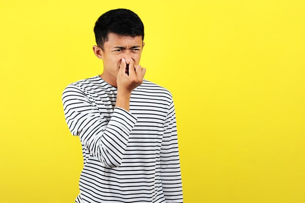Junger asiatischer Mann schließt seine Nase für schlechten Geruch, isoliert auf gelbem Hintergrund