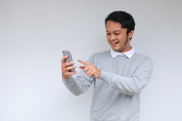 Junger asiatischer Mann schockiert und glücklich über das, was er auf dem Smartphone auf isoliertem grauem Hintergrund sieht