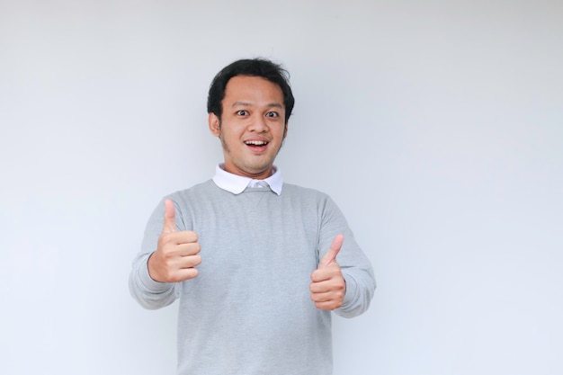 Junger asiatischer Mann trägt graues Hemd mit glücklich lächelndem Gesicht und Daumen hoch oder OK-Zeichen