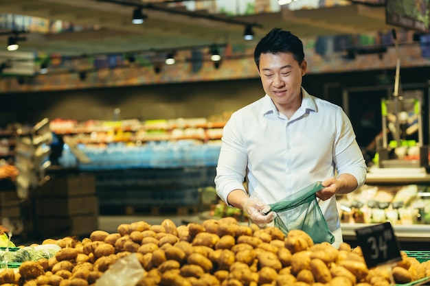 Junger asiatischer mann wählt und pflückt in eco bag gemüse kartoffeln oder obst im supermarkt männlich