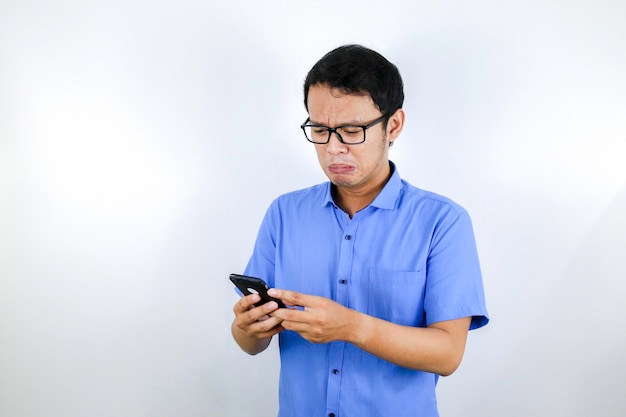 Junger asiatischer Mann weint und ist traurig, wenn er telefoniert und schaut Indonesien Man trägt eine Brille und ein blaues Hemd Isolierter blauer Hintergrund