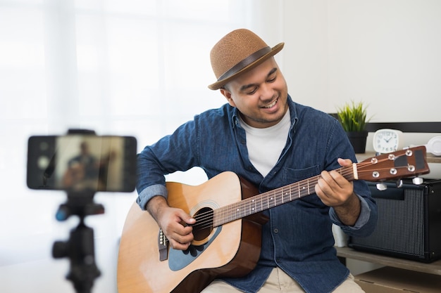 Junger asiatischer Musiker, der die Gitarrenklasse online Live-Streaming mit Smartphone mit Schüler unterrichtet. Indischer Mann, der Akustikgitarre spielt, während er ein Lied singt und per Handy aufnimmt, schattiertes Online-Social