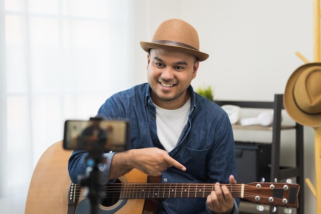 Junger asiatischer Musiker, der die Gitarrenklasse online Live-Streaming mit Smartphone mit Schüler unterrichtet. Indischer Mann, der Akustikgitarre spielt, während er ein Lied singt und per Handy aufnimmt, schattiertes Online-Social