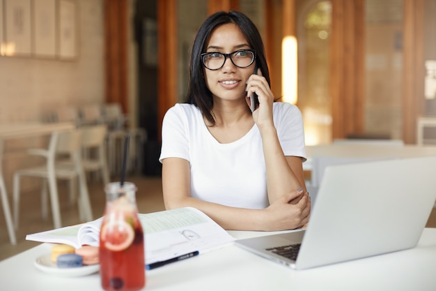 Junger asiatischer Student, der am Telefon eine Brille trägt, die Laptop verwendet, der Limonade in einem hellen, aber leeren Café trinkt