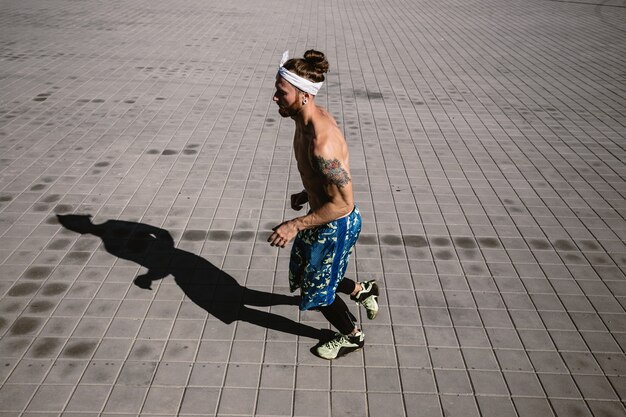 Junger athletischer Mann mit nacktem Oberkörper mit Tätowierungen und Stirnband auf dem Kopf in schwarzen Leggings und blauen Shorts läuft an einem warmen sonnigen Tag auf Pflastersteinen.