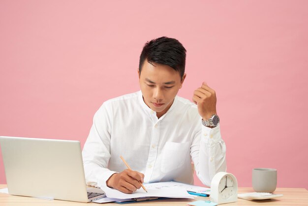 Junger attraktiver asiatischer geschäftsmann, der den laptop verwendet, der auf schreibtischtisch im büro sitzt