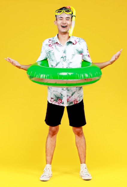 Junger attraktiver asiatischer Mann mit Hut und weißem Hawaiihemd mit gelber Schnorchelmaske und grünem Schwimmring um seine Taille vor gelbem Hintergrund. Konzept für Strandurlaub.