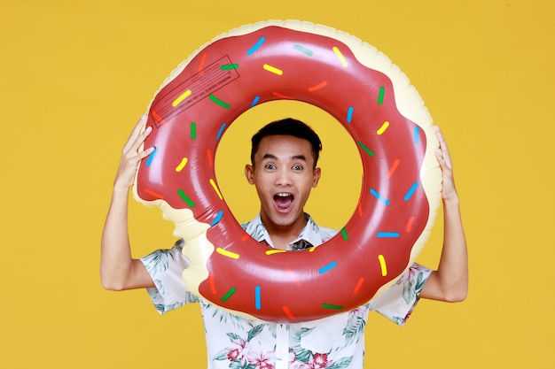 Junger attraktiver asiatischer Mann mit Hut und weißem Hawaiihemd mit roten und grünen Blumenmustern und Donut-Schwimmring vor seinem Gesicht, das Strandurlaub vor gelbem Hintergrund macht.