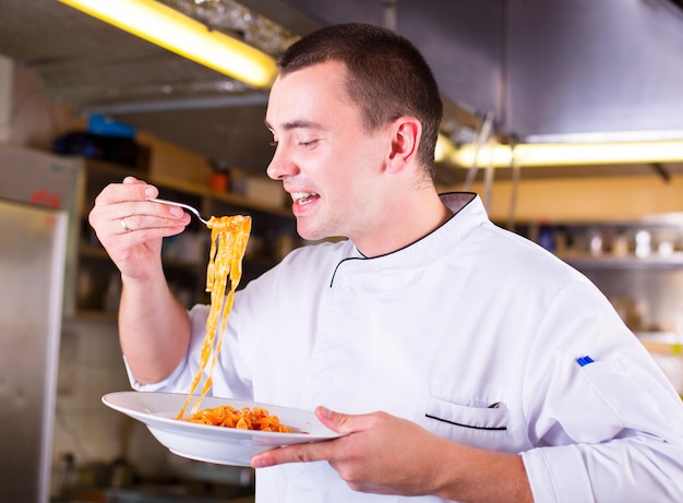 Junger attraktiver Chefmann, der einen Teller hält und die Paste schmeckt. Küchenchef verkostet Essen mit Gabel in der Restaurantküche.