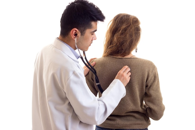 Junger attraktiver Doktor im weißen Kleid mit dem Stethoskop, das dem Rücken der jungen netten Frau zuhört