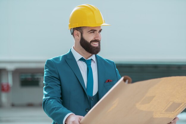 Junger attraktiver erfolgreicher kaukasischer bärtiger Architekt mit gelbem Helm auf Kopf und im blauen Anzug, der Blaupausen hält, während er an Ort und Stelle steht.