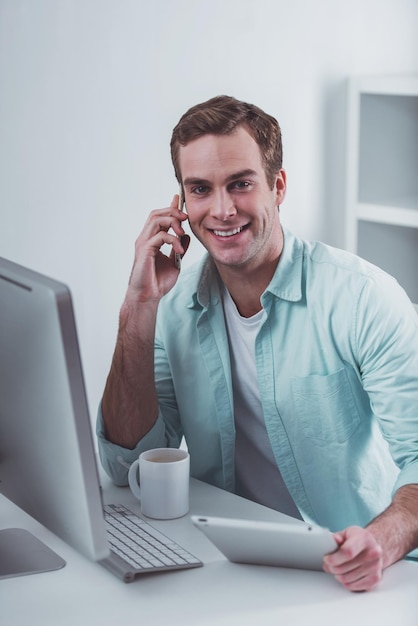 Junger attraktiver Geschäftsmann in Freizeitkleidung mit Tablet, der am Telefon spricht und lächelt, während er im Büro arbeitet