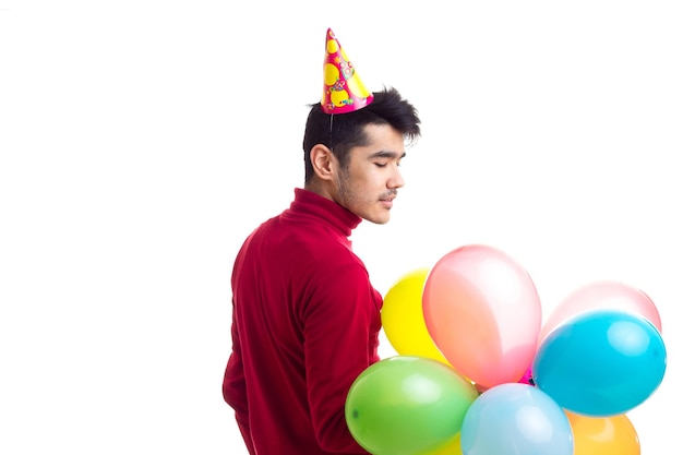 Junger attraktiver Mann im roten Hemd mit feierndem Hut, der viele farbige Luftballons im Studio hält