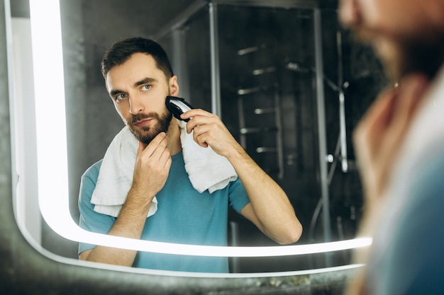 Junger attraktiver Mann mit Handtuch, der den Spiegel betrachtet und sich früh morgens zu Hause rasiert