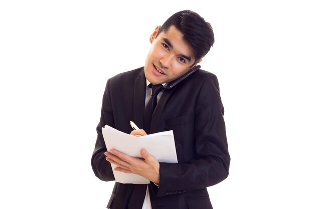 Junger attraktiver Mann mit schwarzen Haaren in Hemd und schwarzem Anzug mit Krawatte, der Papiere hält und redet