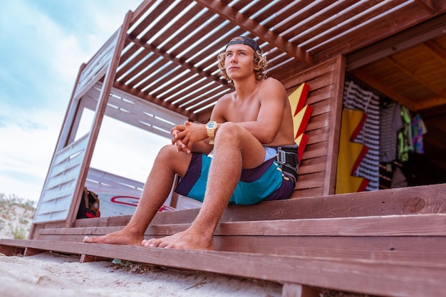 Junger attraktiver Surfer sitzt auf der Terrasse am Strand und blickt in die Ferne zum Meer