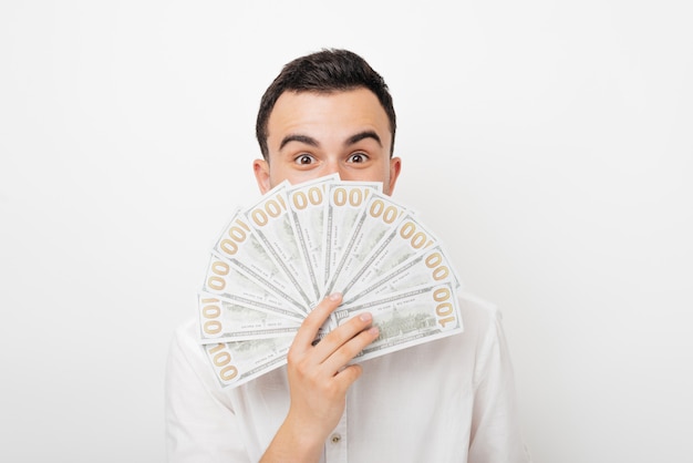 Junger aufgeregter Mann, der sein Gesicht unter den Augen mit einigen Dollars auf weißem Hintergrund bedeckt.