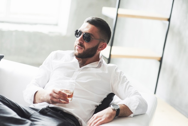 Junger bärtiger Geschäftsmann mit Sonnenbrille und Whisky in der Hand sitzt auf dem weißen Sofa