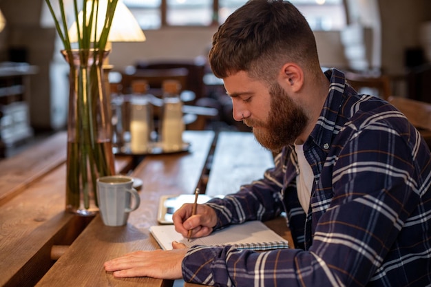 Junger bärtiger Geschäftsmann sitzt im Café, zu Hause am Tisch und schreibt im Notebook, in der Nähe von Lügen-Tablet-Computern. Der Mensch arbeitet, studiert. Online-Bildung, Marketing, Schulung.