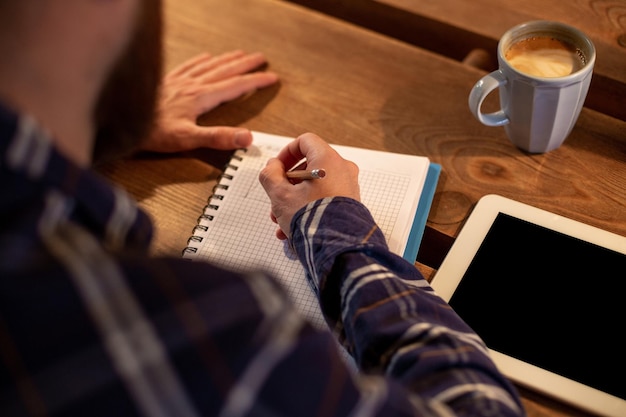 Junger bärtiger Geschäftsmann sitzt im Café zu Hause am Tisch und schreibt in Notebook in der Nähe von Lügen-Tablet-Computer...