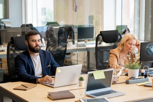 Junger bärtiger Geschäftsmann und reife blonde Frau, die durch Tisch vor Laptops im Büro des offenen Raums sitzen und individuell arbeiten