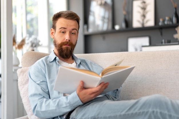 Junger bärtiger gutaussehender Mann liest Buch auf der Couch zu Hause