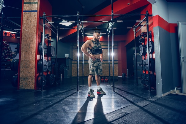 Junger bärtiger kaukasischer ernsthafter Bodybuilder, der im Fitnessstudio mit den Händen auf den Hüften steht.