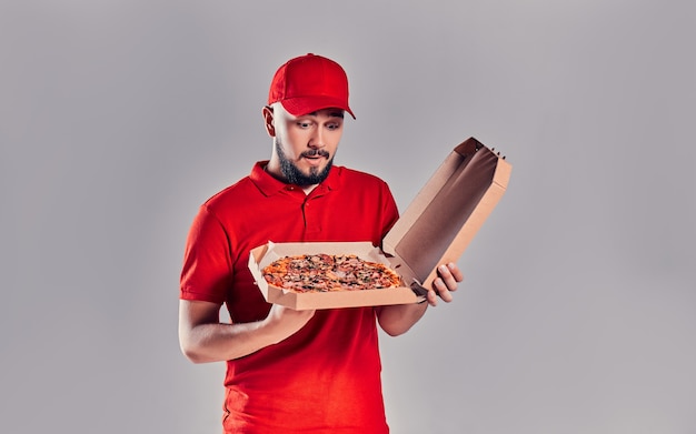 Junger bärtiger Lieferbote in roter Uniform schaut überrascht auf eine Pizzaschachtel, die auf grauem Hintergrund isoliert ist.