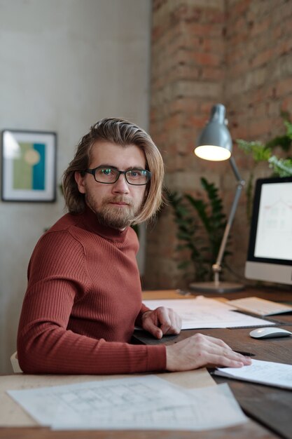 Junger bärtiger männlicher Architekt in Brillen und Freizeitkleidung, der Sie ansieht, während er vor der Kamera am Tisch sitzt und an einem neuen Projekt arbeitet