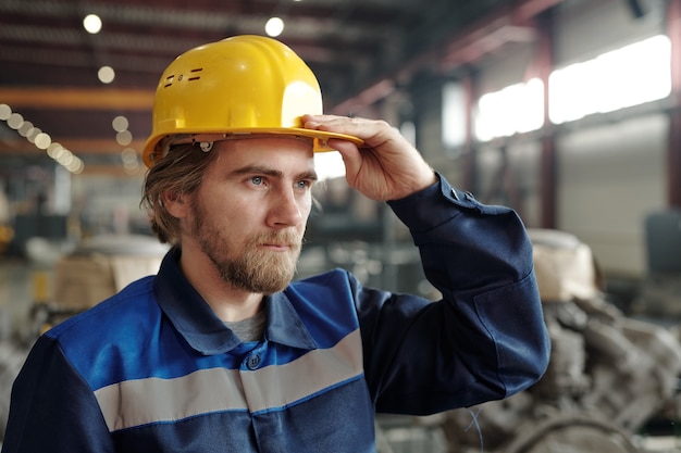 Junger bärtiger männlicher Ingenieur oder Fabrikarbeiter in Schutzhelm, der während der Arbeit in Industrieanlagen vor der Kamera steht