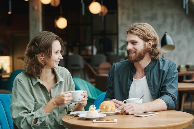 Junger bärtiger Manager sitzt mit Kaffeetasse und Croissant am Tisch und nimmt Sprachnachricht auf