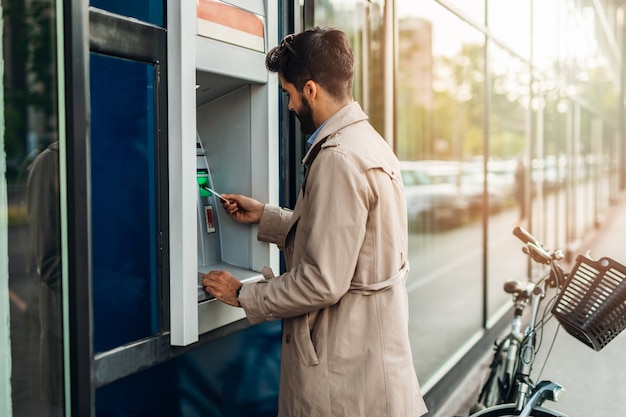 Junger bärtiger Mann, der Geldautomaten verwendet, um Geld abzuheben.